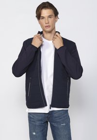 Marineblå pufferjakke med lynlås, tekstureret detalje og sidelommer. Båret over en hvid skjorte, sat sammen med slidte blå jeans.