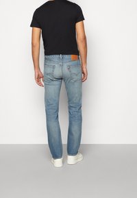 Vaqueros de mezclilla azul claro con corte recto, costuras visibles y una etiqueta de cuero marrón en la parte trasera de la cintura. Combinados con una camiseta negra corta y zapatillas blancas.