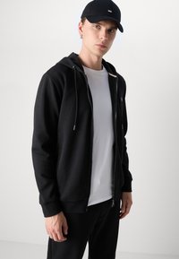 Felpa nera con zip, tasca a marsupio, cappuccio con cordino ed elastici ai polsini, indossata sopra una t-shirt bianca. Accompagnata da un cappellino nero.