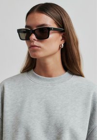 Gafas de sol rectangulares negras con lentes oscuros, con un logo plateado en el lateral, llevadas con un sudadera gris de cuello redondo y textura suave.