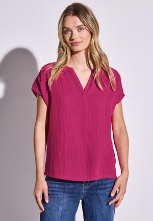 Fuchsia Kurzarmoberteil mit V-Ausschnitt und strukturiertem Stoff, kombiniert mit blauen Jeans. Weiches, fallendes Material mit lockerer Passform.