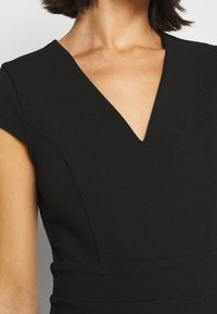 Femme portant une robe noire avec des manches courtes en forme de cape et un décolleté en V, montrant la partie supérieure de la poitrine et la zone des épaules.