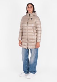 Giacca imbottita beige con cerniera, tasche laterali e cappuccio; presenta un design trapuntato e una texture lucida; abbinata a jeans blu larghi.