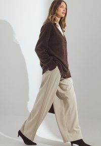 Rudas megztas kardiganas su V formos iškirpte, dviem mygtukais, derinamas su kreminėmis plačiomis kelnėmis ir juodais smailiais kulnais, ant šviesaus fono.