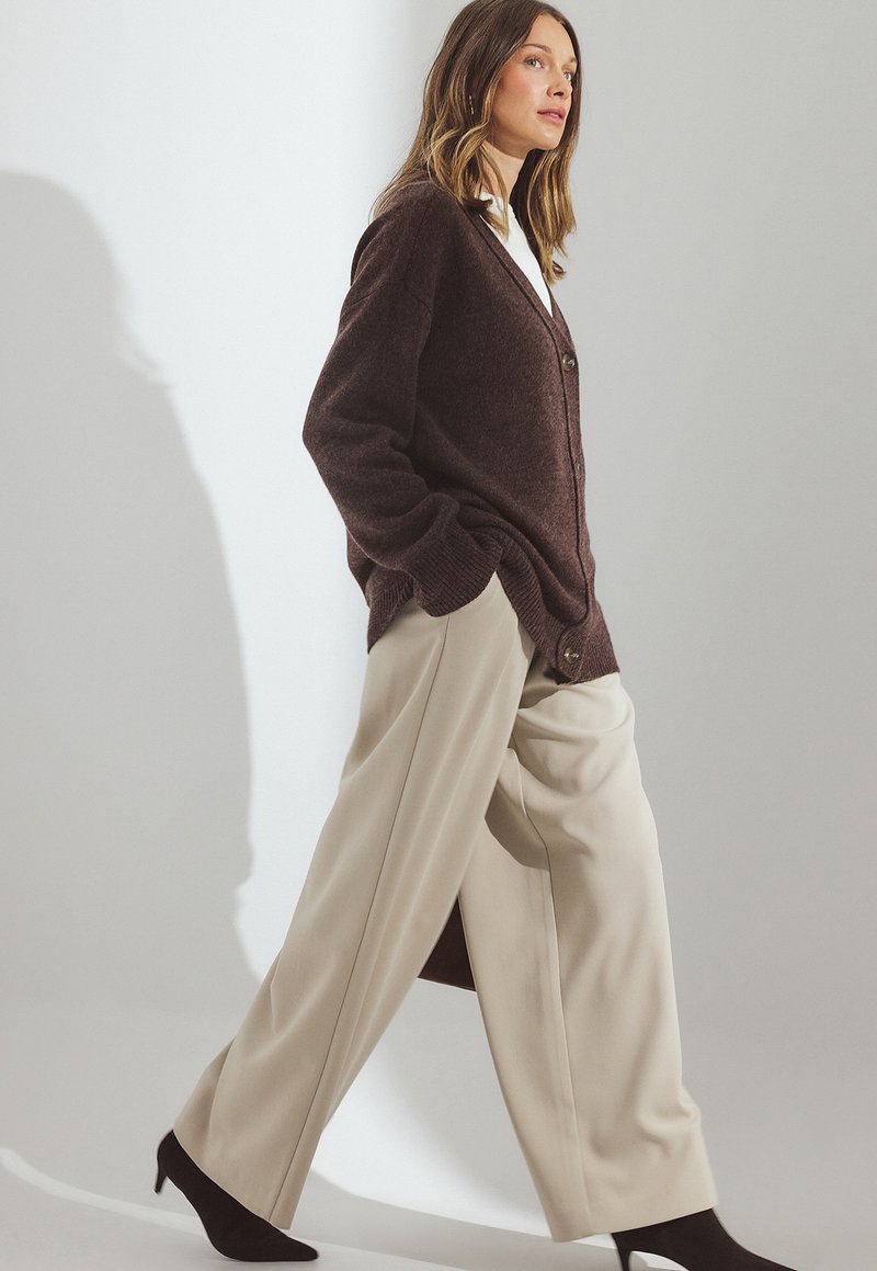 Rudas megztas kardiganas su V formos iškirpte, dviem mygtukais, derinamas su kreminėmis plačiomis kelnėmis ir juodais smailiais kulnais, ant šviesaus fono.
