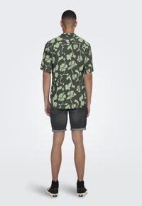 Kurzarmhemd in Dunkelgrün mit einem hellgrünen Blumenmuster; getragen mit hellgrauen, hochgekrempelten Shorts und schwarzen Turnschuhen.