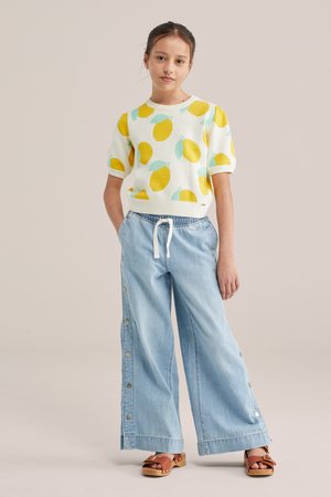 Noor tüdruk seisab käed taskutes, kandes sidrunimustrilise lühikeste varrukatega kampsunit, laia säärega teksapüksid külgnuppudega ja pruune sandaale.