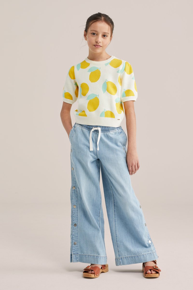 Jong meisje staat met handen in de zakken, draagt een korte mouw trui met citroenprint, wijde denim broek met knoppen aan de zijkant en bruine sandalen.