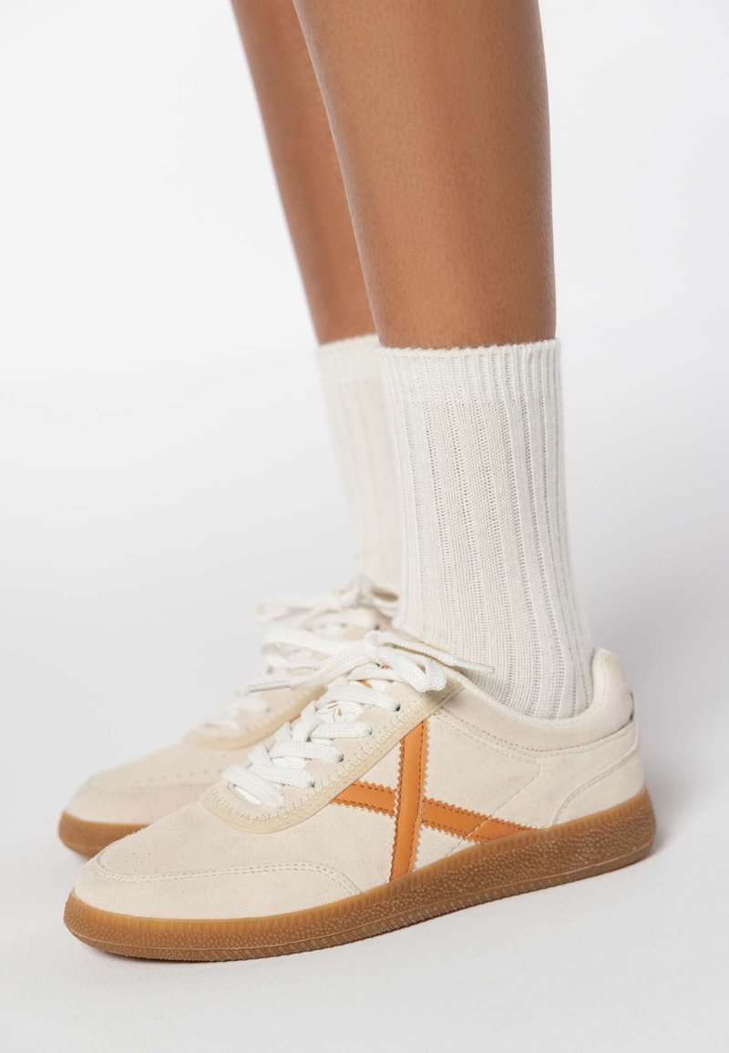 Sneakers in suede beige con accenti arancioni, lacci bianchi e una suola di gomma texturizzata. Indossati con calzini a coste di colore crema.