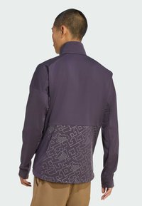 Fioletowa bluza z teksturowaną górną częścią i wzorzystą dolną połową z białymi grafikami. Długie rękawy, wysoki kołnierz i luźny krój.