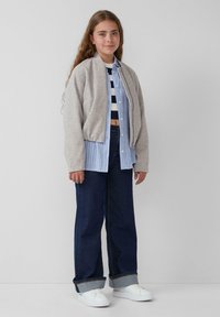 Hellgraue Bomberjacke über einem gestreiften blau-weißen Hemd, kombiniert mit dunkelblauen weiten Jeans und weißen Sneakers. Einfaches Design und Details.