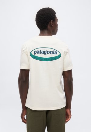 Mężczyzna z krótkimi kręconymi włosami ubrany w kremowy T-shirt Patagonia z zielonym logo i napisem, w połączeniu z oliwkowozielonymi spodniami, zwrócony tyłem.