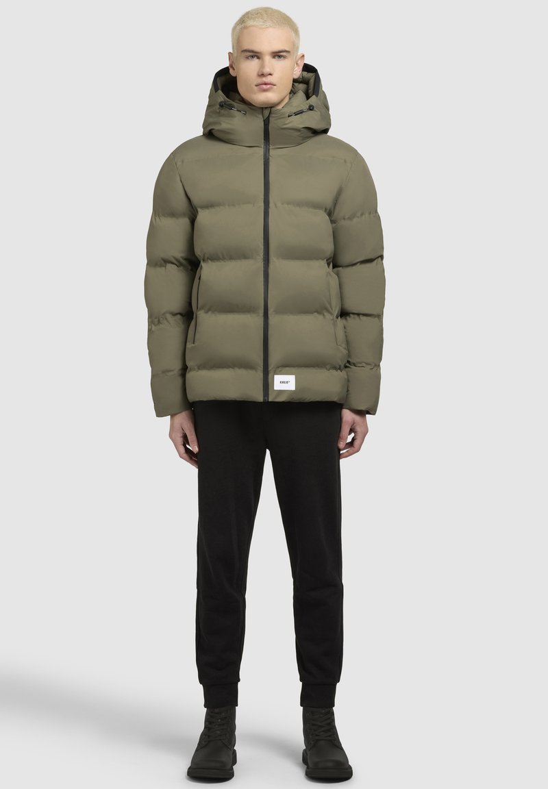 Zaļa pūkaina jaka ar kapuci, ar rāvējslēdzēju un kontrastējošu melnu rāvējslēdzēju. Uz apakšējās apmales ir uzlīme, jaka valkāta virs melnām biksēm un botēm.