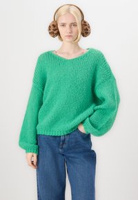 Grøn strikket sweater med v-hals, oversized ærmer og struktureret stof, parret med blå højtaljede jeans og brune ørevarmere med leopardmønster.