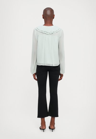 Light green, long-sleeve blouse with ruffled collar detail, sheer fabric, fitted waist, paired with black flared pants and open-heel shoes.