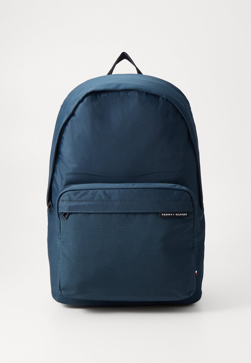 Sac à dos Tommy Hilfiger bleu marine avec une poche avant zippée et une poignée supérieure noire sur fond blanc.