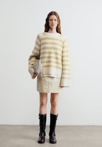 Γυναίκα που στέκεται σε εσωτερικό χώρο, φορώντας πουλόβερ με μπεζ και λευκές ριγέ, κρεμ φούστα, μαύρες μπότες μέχρι το γόνατο, κρατώντας μια μπεζ τσάντα φάκελο.