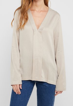 Femme portant un chemisier beige à manches longues avec un col en V et une coupe ample, associé à un jean bleu, debout devant un fond clair uni.
