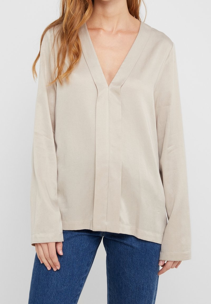 Femme portant un chemisier beige à manches longues avec un col en V et une coupe ample, associé à un jean bleu, debout devant un fond clair uni.