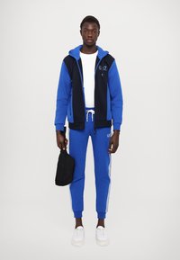 Completo sportivo blu con giacca con zip e pantaloni abbinati. Presenta accenti bianchi e un logo, abbinati a sneakers bianche e una borsa nera.