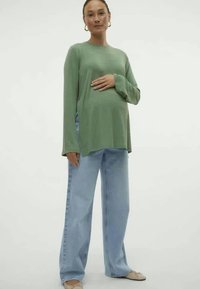 Zeleni dolgi rokav s sproščenim krojem v kombinaciji z svetlo modrimi širokimi hlačami in nevtralnimi balerinkami, kar ustvarja sproščen in udoben videz.