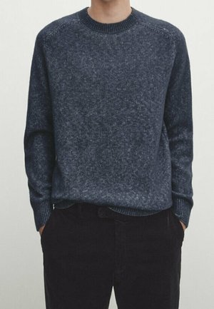 Personne portant un pull en maille bleu foncé à col rond et un pantalon noir, les mains dans les poches, devant un fond clair uni.