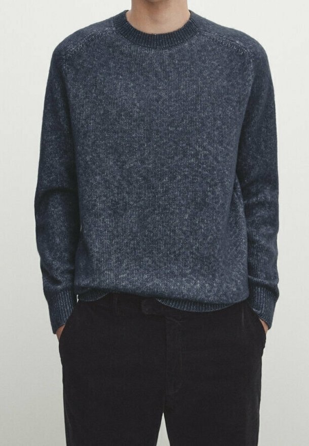 Personne portant un pull en maille bleu foncé à col rond et un pantalon noir, les mains dans les poches, devant un fond clair uni.