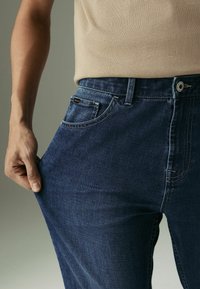 Blugi din denim albastru închis, cu un design drept, având buzunare frontale, închidere cu nasture și o țesătură texturată. O mână strângând talia.