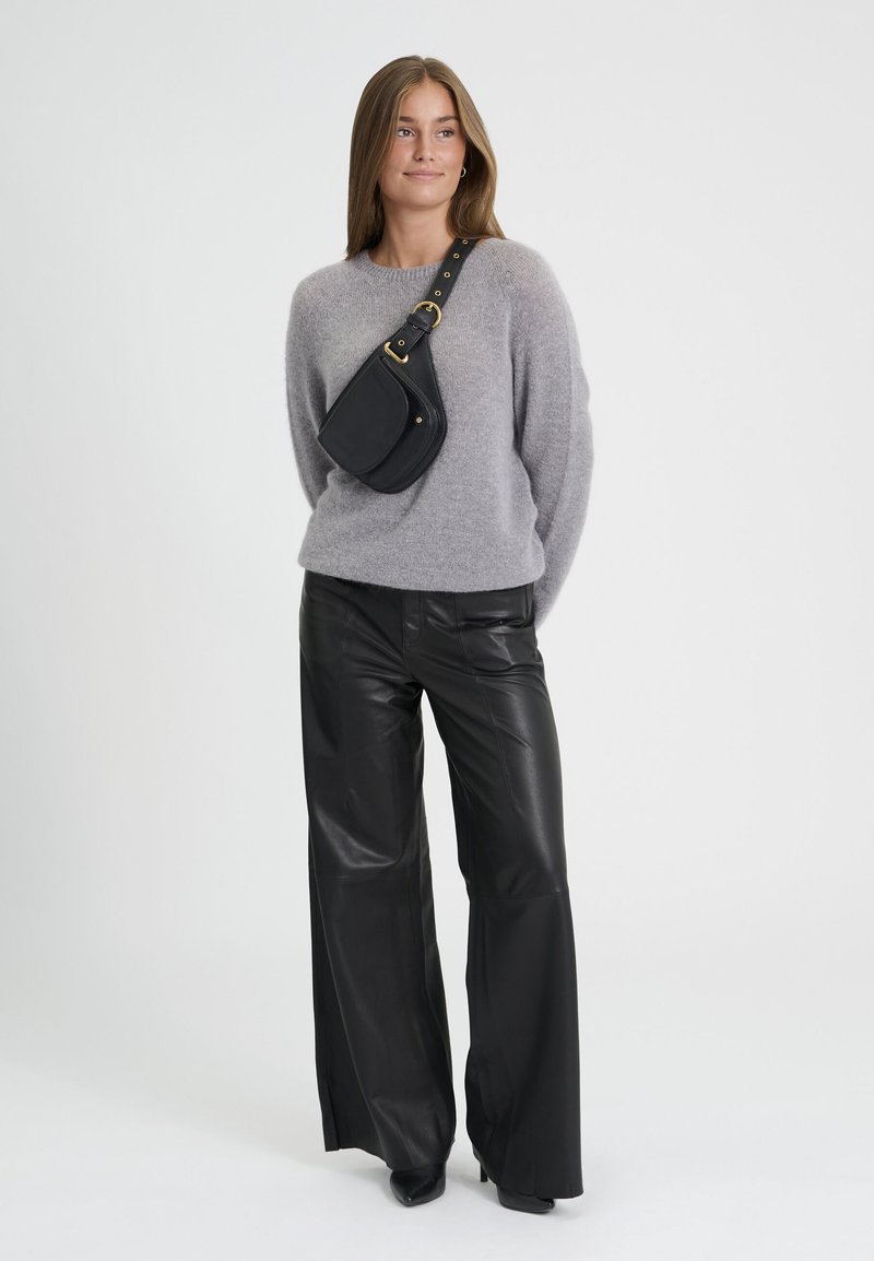 Jeune femme portant un pull gris, un pantalon large en cuir noir, un sac bandoulière noir et des chaussures noires, debout devant un fond uni.