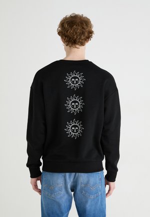 Schwarzer Sweatshirt aus Baumwolle mit weißen Sonnenmotiven auf der Rückseite. Rippbündchen und Saum. Kombiniert mit hellblauen Jeans.