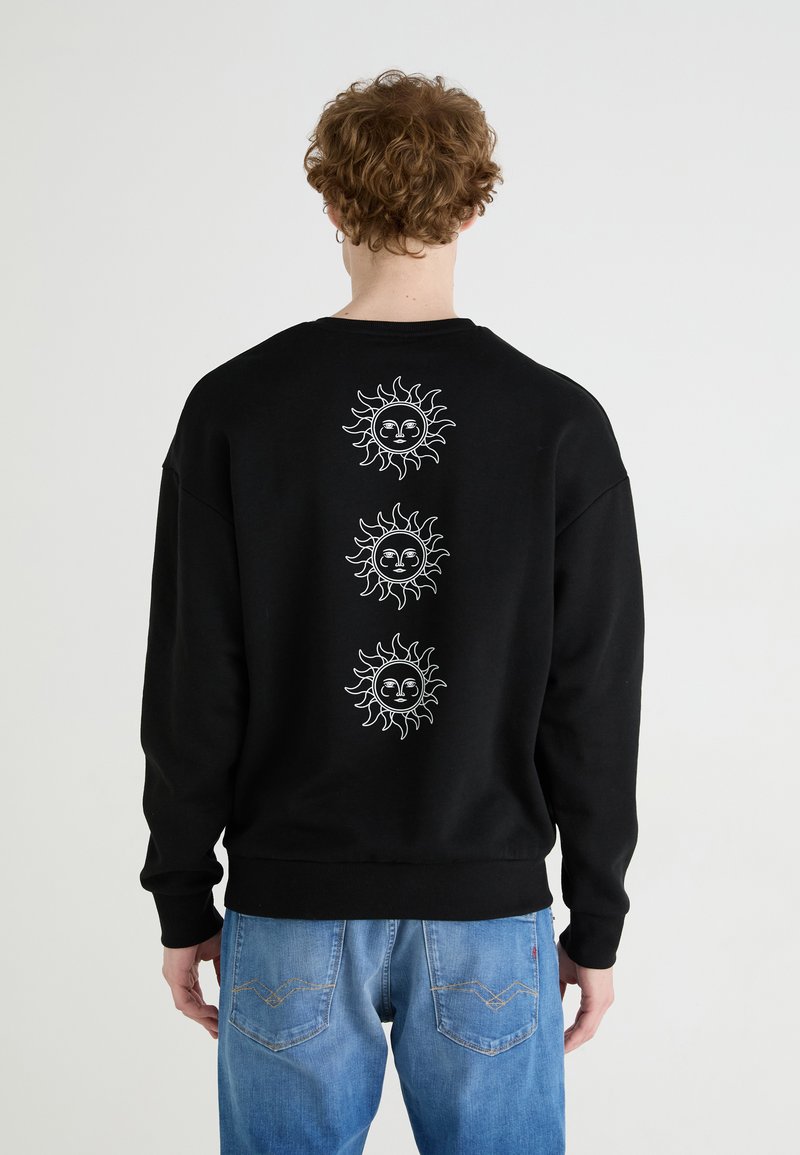 Felpa nera in cotone, con grafica di sole bianco sulla schiena. Cuffs e orlo a coste. Abbinata a jeans blu chiaro.