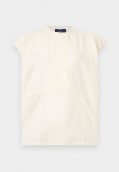 Κρεμ μπλούζα με κοντά μανίκια, στρογγυλή λαιμόκοψη, τρία κουμπιά μπροστά και διακριτικές πιέτες κοντά στους ώμους και τη μέση.