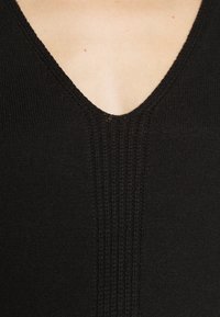 Gros plan d’un vêtement en maille noire avec un décolleté en V montrant une partie de la peau nue et de petites taches près de la clavicule.