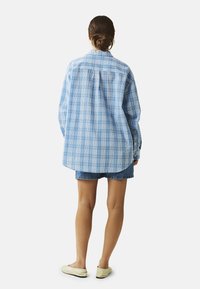 Světle modrá kostkovaná košile volného střihu s špičatým límcem, knoflíkovými manžetami a zakřiveným lemováním, v kombinaci s džínsovými šortkami a světlými botami.