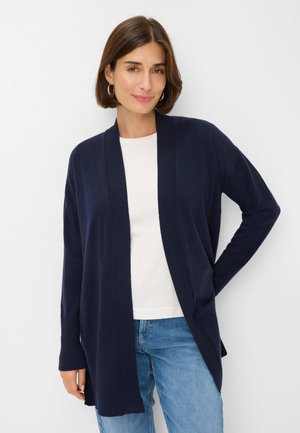 Marineblauwe cardigan gemaakt van zachte brei stof, open voorkant, geribbelde manchetten en zoom, gecombineerd met een wit shirt en lichtblauwe jeans.