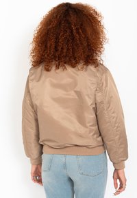 Person mit lockigem roten Haar, der eine beige Bomberjacke und hellblaue Jeans trägt, steht mit dem Rücken zur Kamera vor einem weißen Hintergrund.