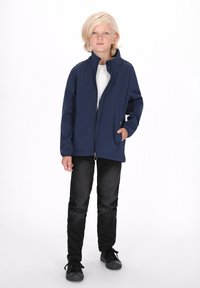Jachetă din softshell bleumarin cu guler înalt, fermoar complet și buzunar lateral, purtată cu blugi negri și adidași negri. Material gros, texturat.