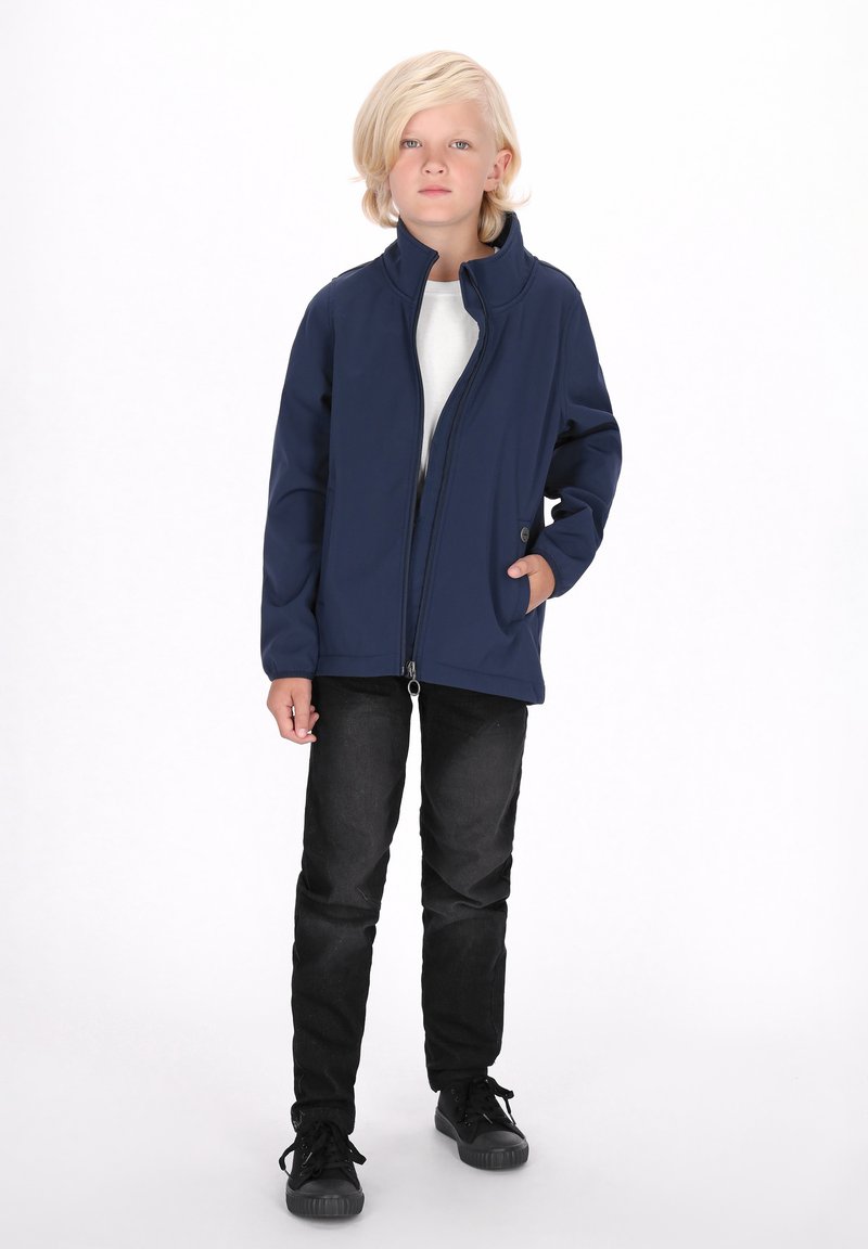 Jachetă din softshell bleumarin cu guler înalt, fermoar complet și buzunar lateral, purtată cu blugi negri și adidași negri. Material gros, texturat.