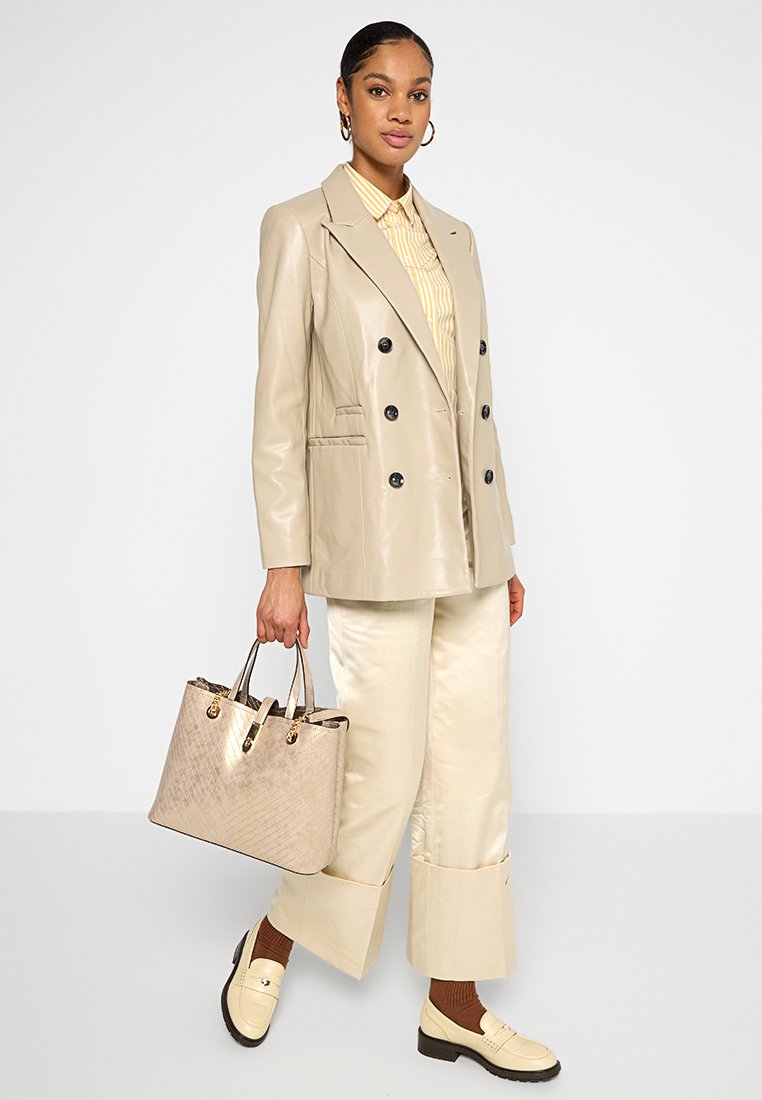 Blazer beige de cuero sintético de doble botonadura, camisa a rayas amarillas, pantalones de satén de pierna ancha y un bolso texturizado de color marrón claro. Mocasines crema completan el look.