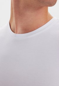 Hvid T-shirt med rund hals lavet af glat, strækbart bomuld. Nærbillede viser halsudskæringen og den bløde tekstur, der fremhæver de minimale sømme og pasformen.