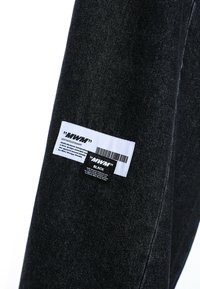 Chaqueta de mezclilla negra con una etiqueta de cuidado blanca con texto negro, código de barras y logo de la marca en la manga izquierda. Textura suave, diseño casual.