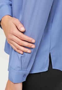 Camisa de manga larga azul claro con una textura suave, puños abotonados y aberturas laterales. La mano reposa suavemente sobre la manga.