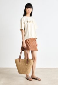 Gai beige lielizmēra t-krekls ar zelta logotipu, brūnas ādas mini svārki, austs salmu soma ar brūnām rokturām un teksturētas pelēkas slaidu sandales.