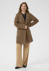 Cappotto in misto lana marrone con collo a rovescio, chiusura con bottoni frontali e due tasche laterali, abbinato a pantaloni beige a gamba larga e scarpe nere.