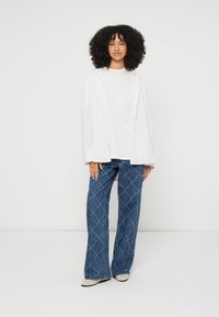 Casaco oversized branco com riscas, t-shirt branca lisa e jeans azuis com padrão geométrico em forma de diamante. Sapatos bege, posicionado contra um fundo branco.