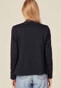 Blazer navy con textura sutilmente estampada, ajuste entallado, mangas largas y cuello estructurado, combinado con jeans de color azul claro.