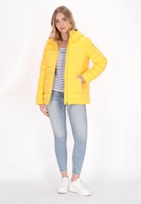 Giacca imbottita gialla con zip frontale e cappuccio, abbinata a una maglietta a righe e jeans skinny azzurri, completata da sneakers bianche.