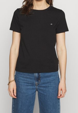 Mujer con camiseta negra de manga corta de Calvin Klein y vaqueros azules, con una pulsera dorada en la muñeca izquierda.