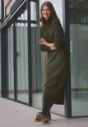 Olivgrünes, strukturiertes, gestricktes Midi-Kleid mit hohem Kragen und langen Ärmeln. Getragen mit schwarzen knielangen Stiefeln und einer Holz-Plateausohle.