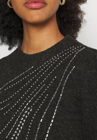 Dunkel gestrickter Pullover mit rundem Ausschnitt, verziert mit einem Muster aus silbernen Perlen und rechteckigen Pailletten in einem diagonalen Design.